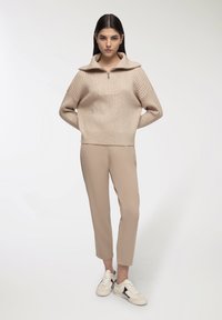 Beżowy sweter prążkowany z wysokim kołnierzem i zamkiem, w połączeniu z beżowymi spodniami typu tapered i białymi trampkami z czarnymi akcentami.