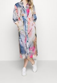 Kobieta ubrana w kolorową sukienkę midi z efektem tie-dye, z rękawami 3/4, regulowanym paskiem w talii, kieszeniami i białymi trampkami, stojąca na białym tle.