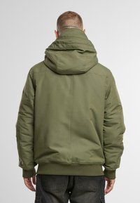 Chaqueta acolchada de color verde oliva con capucha, puños y bajo acanalados, y textura suave. Presenta costuras minimalistas y bolsillos laterales.
