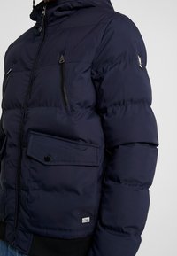 Marine pufferjack met capuchon, voorzien van ritszakken, een drukknopflapzak en ribgebreide boorden. Gladde textuur met horizontale naden.