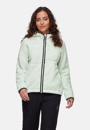 Frau mit lockigem Haar, die eine hellgrüne Fleecejacke mit Kapuze und schwarze Hose trägt, steht vor einem einfarbigen Hintergrund.