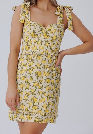 Ærmeløs cremefarvet kjole med gult citron- og grønt bladprint, skuldre med bindebånd, flæsekant ved talje og nederkant, båret af en stående person.