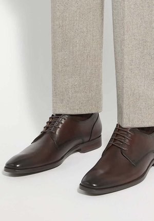 Chaussures habillées en cuir marron foncé avec lacets portées avec un pantalon beige clair, montrées de la moitié du mollet vers le bas sur un fond uni.