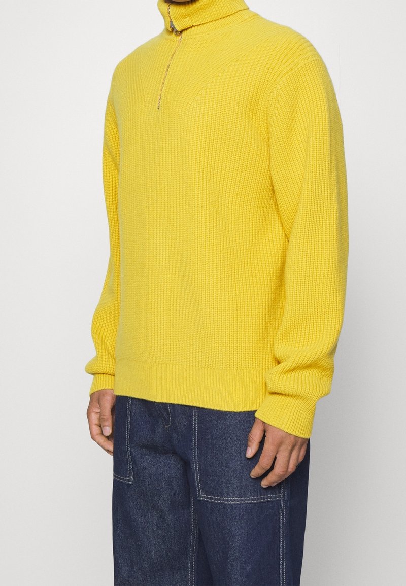 Geel gebreid sweater met een hoge kraag en halve rits. Kenmerkt zich door een geribbelde textuur en lange mouwen. Gecombineerd met donkerblauwe spijkerbroek.