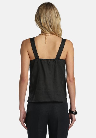 Femme blonde portant un haut noir sans manches à larges bretelles et un pantalon noir, vue de dos sur un fond uni.