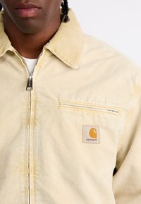 Veste en velours côtelé beige clair avec fermeture éclair, col pointu et une poche avant arborant un logo Carhartt. Matériau texturé.