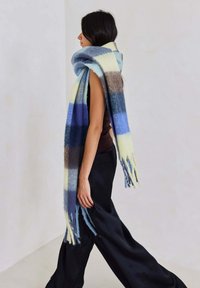 Wollen plaid sjaal met blauwe, gele, grijze en bruine strepen; zachte textuur met franjes aan de uiteinden; om de hals gewikkeld.