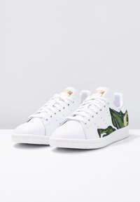 Valkoiset nahkasneakerit, joissa on kirkkaanvihreät lehtikuvioinnit sivuilla, tasaiset nauhat ja kultaiset yksityiskohdat kielen kohdalla.