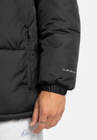 Crna puffa jakna s zatvorenim džepom, elastičnim rukavima i detaljem logotipa na rukavu, izrađena od glatkog i izdržljivog materijala.