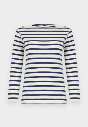 Prúžkované tričko s dlhým rukávom v bielej a námorníckej modrej, doplnené širokými modrými džínsami. Modelka drží tmavozelenú semišovú kabelku so zlatými kovaniami.