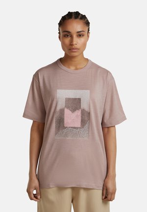 Woman with braided hair wearing a loose mauve t-shirt with abstract geometric design and beige pants, standing against a plain background.