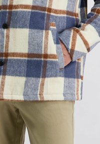 Man in een blauw, bruin en wit geruit jasje met zijn handen in de zakken, gecombineerd met beige broeken.