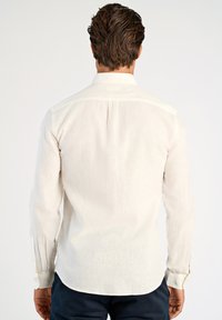 Camicia beige chiaro a maniche lunghe con collo classico, schiena dritta e polsini con bottoni. Tessuto morbido e strutturato con un dettaglio di trama sottile.