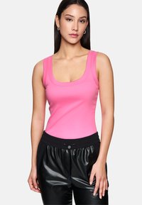 Woman wearing pink sleeveless top and black faux leather pants, standing against plain light background with neutral expression.