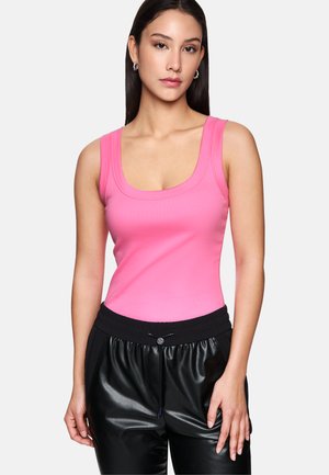 Frau in pinkem ärmellosem Top und schwarzen Kunstlederhosen, steht vor einem schlichten hellen Hintergrund mit neutralem Gesichtsausdruck.