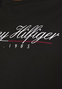 Černé bavlněné tričko s bílým kurzívním nápisem „Tommy Hilfiger“ a červenou vodorovnou čarou nad číslem „1985“. Hladká textura, kulatý výstřih.