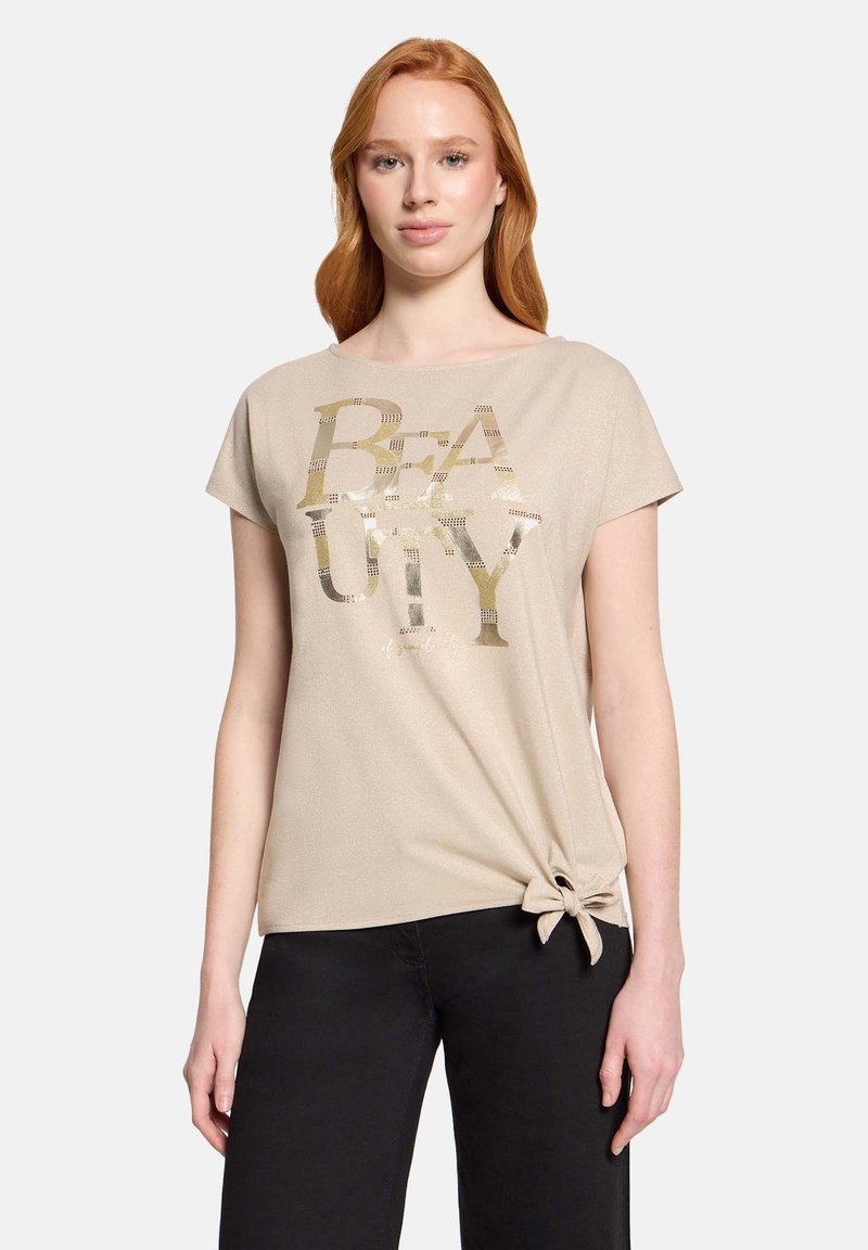 Femme aux longs cheveux roux portant un haut beige à manches courtes avec l'inscription métallique "BEAUTY" et un nœud sur le côté, associée à un pantalon noir, debout devant un fond blanc.