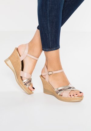 Fødder iført beige kilehæls-sandaler med ankelremme og metalliske detaljer, kombineret med mørkeblå skinny jeans på hvid baggrund.