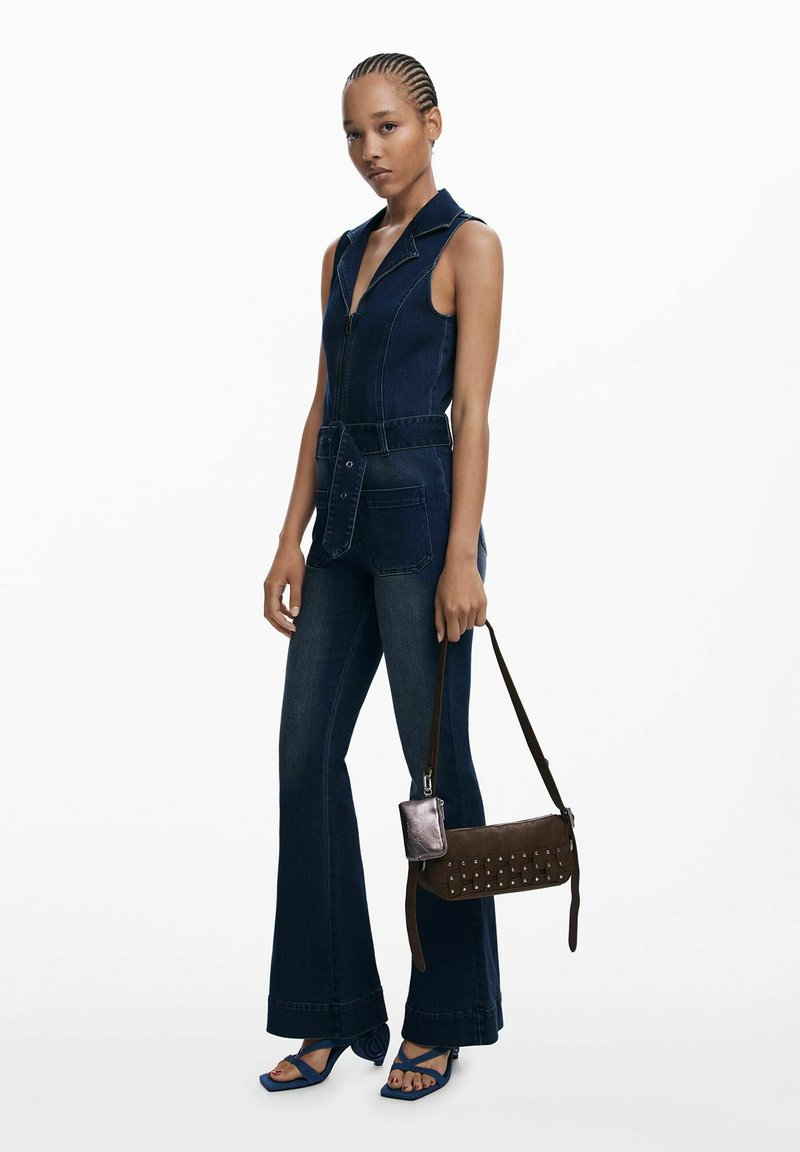 Der dunkelblaue Jeansoverall verfügt über einen Reißverschluss vorne, Taschen und eine taillierte Silhouette mit Gürtel. Die Accessoires umfassen eine kleine braune Tasche und blaue Sandalen.