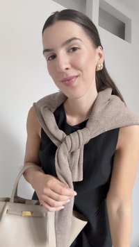 Beige ribbed sweater draped around the shoulders, black dress, holding a beige handbag with a gold clasp, smooth texture and structured shape.