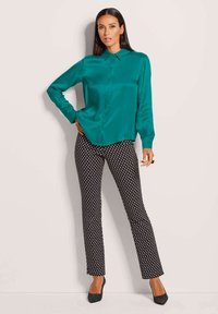 Vrouw die met één hand op de heup staat, gekleed in een teal satin blouse met lange mouwen, zwart-witte broek met patroon en zwarte hakken.