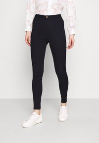 Leggings negras con una textura suave, cintura alta y un detalle de botón dorado. Combinados con zapatillas blancas para un look casual.