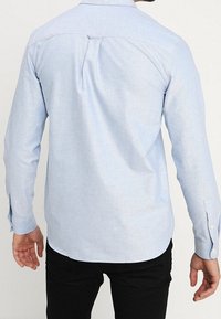 Camisa de manga larga azul claro con un pliegue en la espalda, puños abotonados y un corte entallado, confeccionada en una tela suave y texturizada.