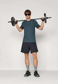 Conjunto deportivo en negro y gris que incluye una camiseta ajustada en teal oscuro, pantalones cortos negros y zapatillas negras. Sosteniendo una barra, lleva puestas gafas de sol.