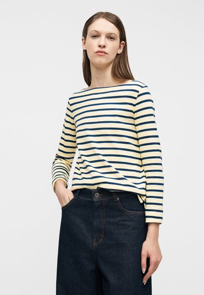 Γυναίκα με καστανά ίσια μαλλιά που φοράει μακρυμάνικη μπλούζα με κρεμ και ναυτικό μπλε ρίγες και ψηλόμεσο σκούρο μπλε τζιν, ποζάρει με το ένα χέρι στην τσέπη.