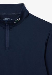 Haut à manches longues bleu marine avec un col à fermeture éclair au quart, fabriqué en tissu lisse. Présente un petit logo Lacoste vert sur le col.
