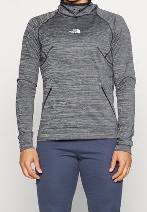 Personne portant un pull-over ajusté gris chiné à manches longues avec un col montant et le logo The North Face, associé à un pantalon foncé.