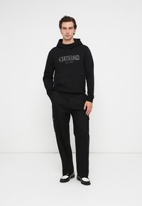 Czarna bluza z kapturem z logo "GUESS", z przednią kieszenią, zestawiona z czarnymi spodniami cargo z bocznymi kieszeniami i czarno-białymi butami.