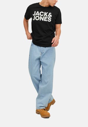 Man wearing black "JACK & JONES" t-shirt, light blue loose jeans, and tan work boots, standing with one leg crossed in front.