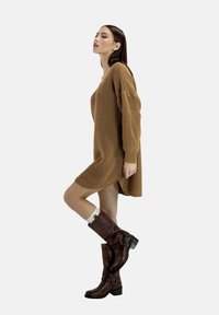 Brown knitted sweater dress with long sleeves and side slits, paired with knee-high brown leather boots featuring buckle accents.