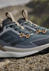 Een paar donkergrijze en beige trailrunning schoenen met gestructureerde zolen die rusten op rotsachtige grond buiten.