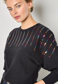 Pull noir à col rond avec des rayures de sequins colorés sur la poitrine en rose, bleu, or et cuivre. Texture douce.