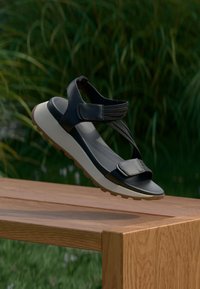 Sandale sportive noire avec des brides ajustables et une épaisse semelle blanche, posée sur un banc en bois en plein air avec un fond de feuillage vert.