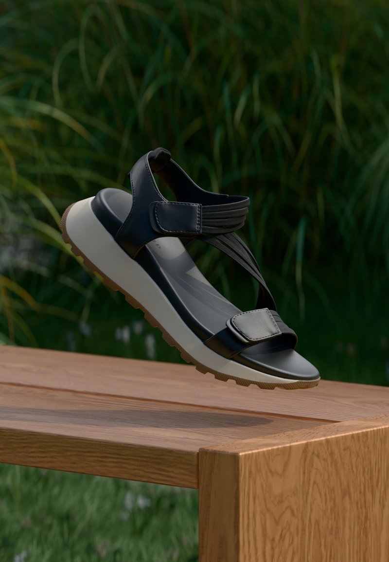 Sandale sportive noire avec des brides ajustables et une épaisse semelle blanche, posée sur un banc en bois en plein air avec un fond de feuillage vert.