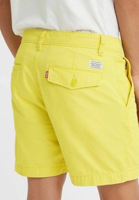 Gula bomullsshorts med en slät textur, med bakfickor, komfortknäppning och en liten märkslapp synlig på midjebandet.