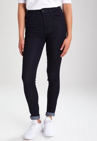 Vaqueros de corte alto de lavado oscuro con un ajuste ceñido. El tejido es de denim suave, con un corte recto y puños enrollados, combinados con zapatillas blancas.
