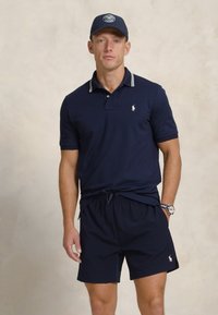 Tricou polo bleumarin cu guler cu dungi albe, asortat cu pantaloni scurți bleumarin. Ambele articole au accente cu logo. Include un ceas și o șapcă.