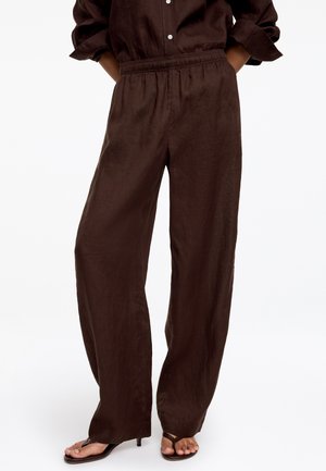 Vrouw die losse donkerbruine linnen broek met elastische taille en opgerolde donkerbruine blouse draagt, gecombineerd met open teen sandalen met hak.