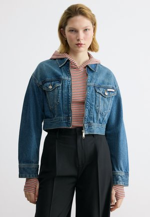 Νεαρή γυναίκα που φοράει ένα κοντό τζιν μπουφάν σε μπλε χρώμα πάνω από μια κόκκινη και λευκή ριγέ μπλούζα με κουκούλα, συνδυασμένο με μαύρο ψηλόμεσο παντελόνι.