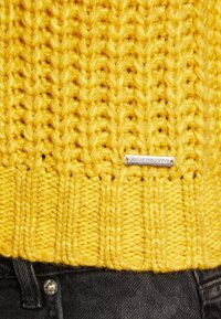 Geel gebreide trui met een textuurpatroon, voorzien van een geribde zoom. Een zilveren label met de tekst "COLOURFUL REBEL" staat op de voorkant.
