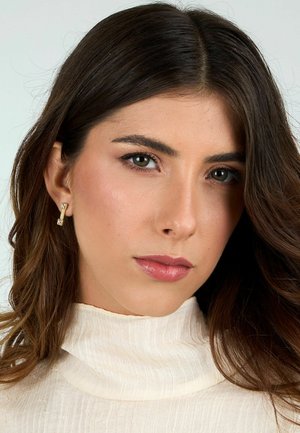 Orecchini color oro con accenti rettangolari e quadrati, indossati con un top a collo alto di colore crema e texture. La pelle liscia e i capelli scuri e mossi incorniciano il viso.
