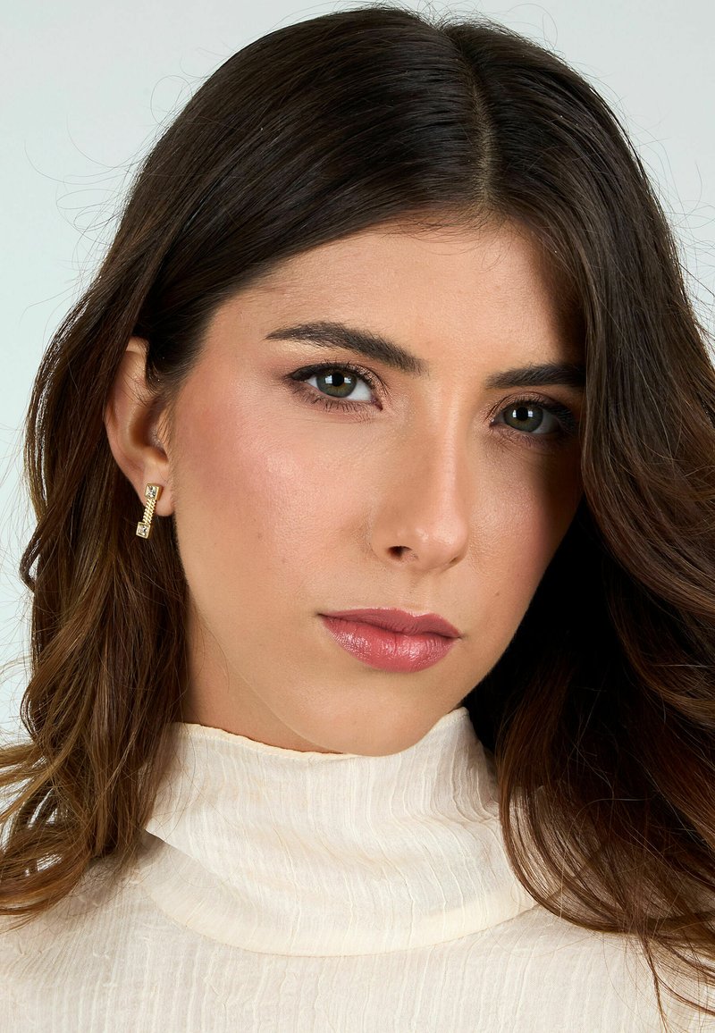 Orecchini color oro con accenti rettangolari e quadrati, indossati con un top a collo alto di colore crema e texture. La pelle liscia e i capelli scuri e mossi incorniciano il viso.