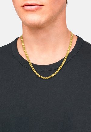 Collana in metallo dorato con un design intrecciato, indossata sopra una maglietta nera a maniche corte con scollo rotondo.