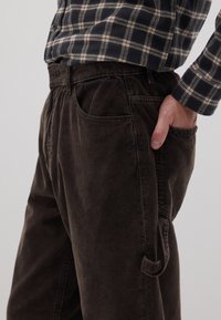 Bruine corduroy broek met een ontspannen pasvorm, voorzien van een zijzak en getextureerde ribbels. Draagt met een geruiten overhemd in marineblauw en beige.