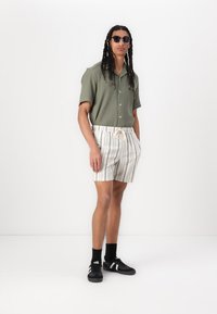 T-shirt vert à manches courtes en côte, shorts rayés beige et blanc avec cordon de serrage, baskets noires et lunettes de soleil. Texture lisse, coupe décontractée.