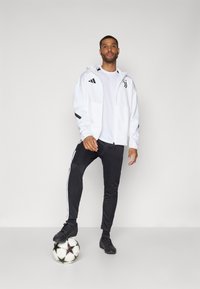 Hvid zip-up jakke med sorte detaljer, parret med sorte joggers med hvide striber. Sort fodbold ved fødderne, stående stilling.